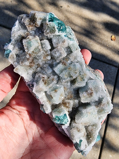 In front of a blurred outdoor background, someone holds an enchanting Fluorite with Quartz cluster from the Druzy Dreams Pocket at Rogerley Mine, featuring green and whitish cubic crystals. The surface is textured and sparkly, showcasing the artistry of The Crystalary's specimen, sku11.