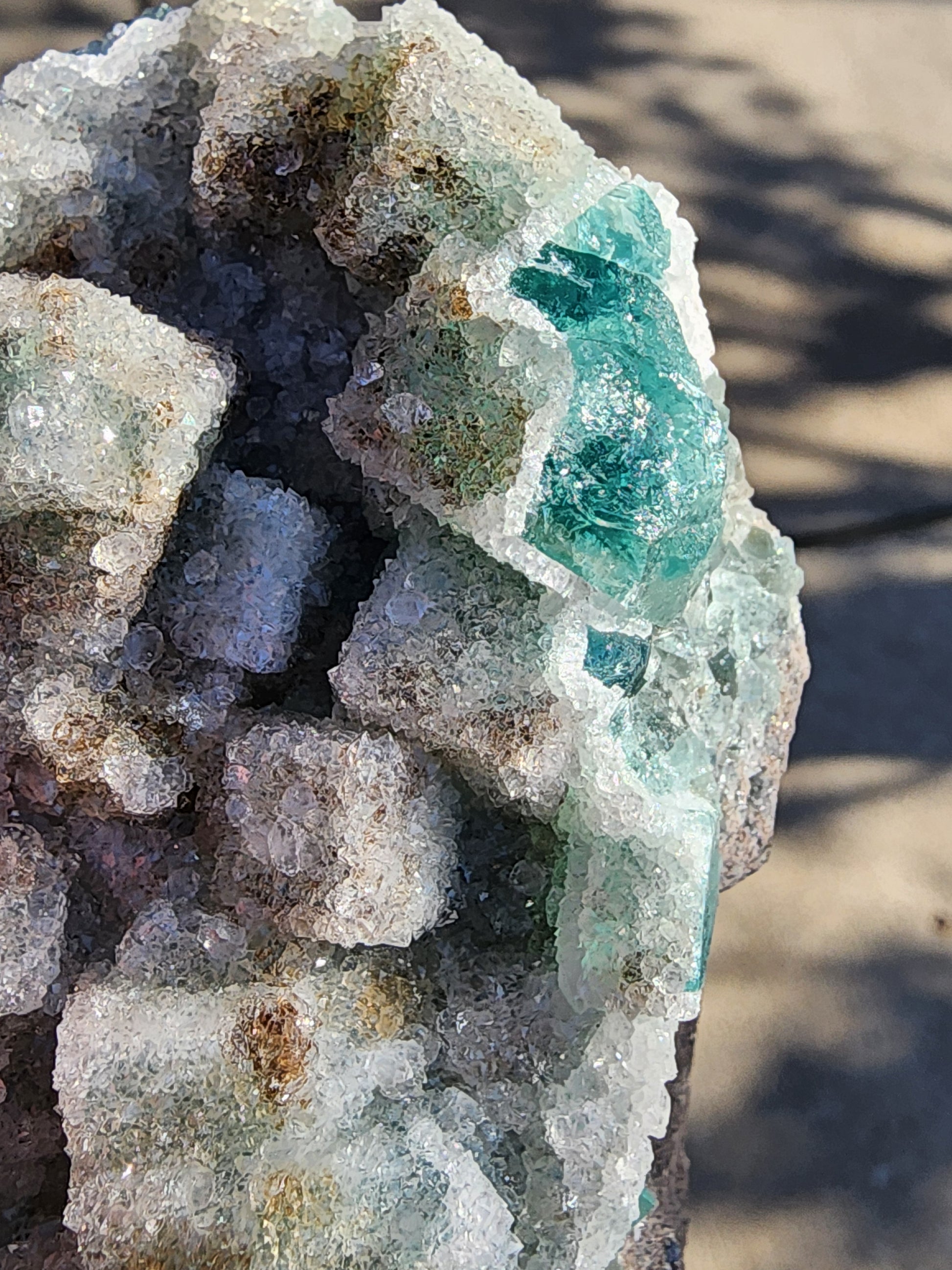 A close-up of the "Fluorite with Quartz - Druzy Dreams Pocket" from The Crystalary reveals clear and green crystals, likely sourced from Rogerley Mine. The jagged cluster SKU11 specimen's textured formations glisten in the sunlight, set against a blurred outdoor background that enhances its natural sparkle and intricate beauty.