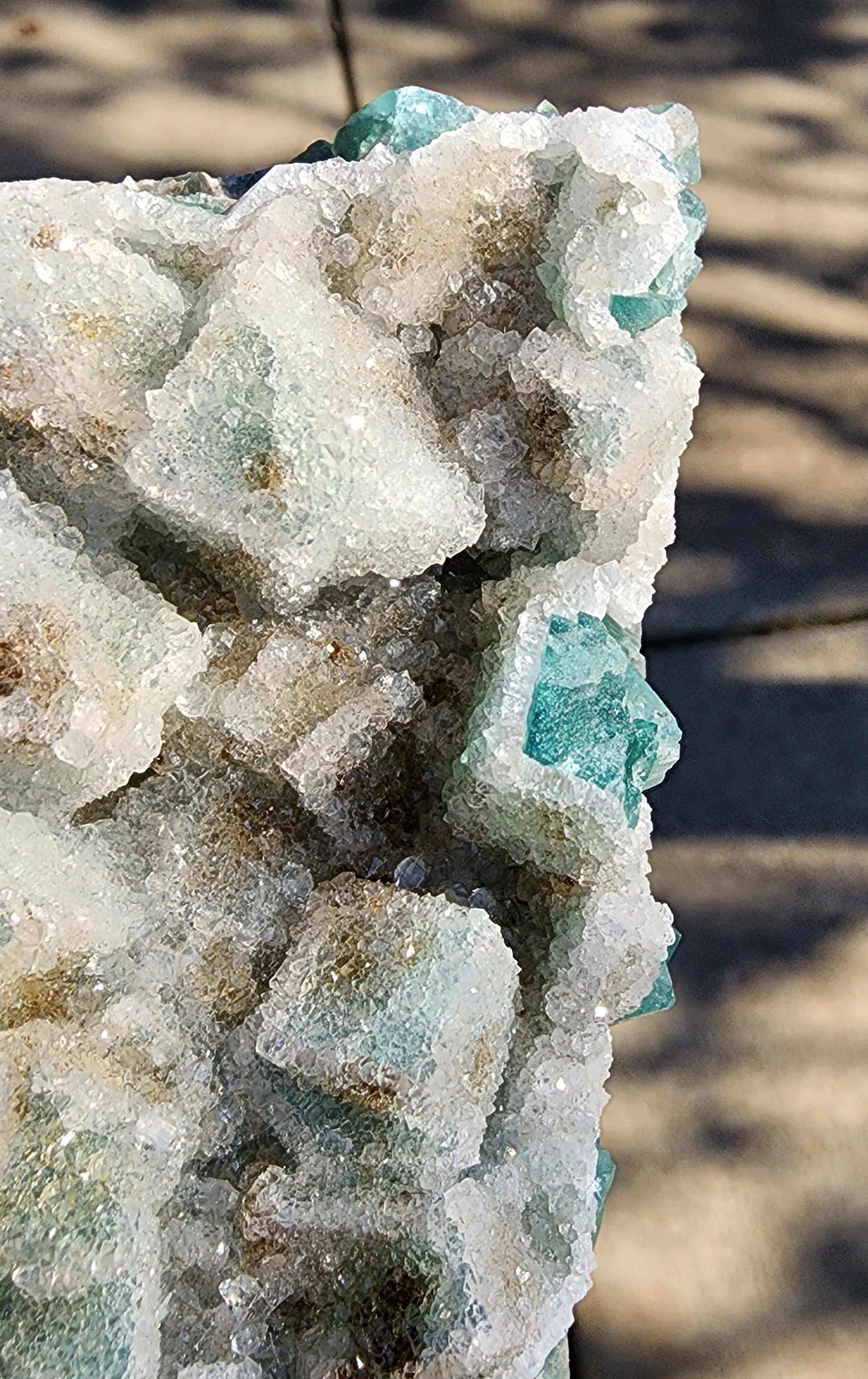 A close-up of the Fluorite with Quartz- Druzy Dreams Pocket from Rogerley Mine, offered by The Crystalary, showcases its green and white crystalline structures. Sunlight casts shadows on the uneven surfaces, accentuating the texture and sparkle of this exquisite fluorite cluster specimen. In the background, a blurred sidewalk further enhances its beauty.