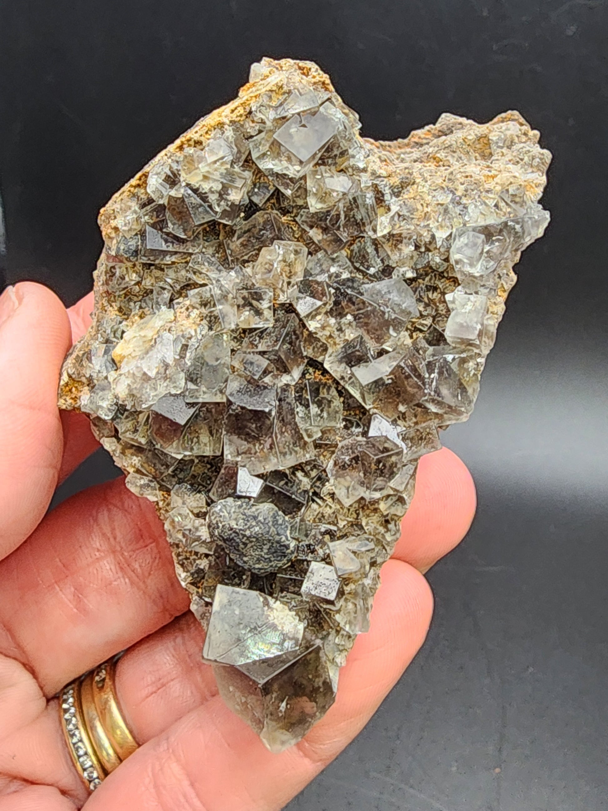 A hand holds a rough, translucent cluster of Fluorite from the Smoky Rabbit Pocket at the Diana Maria Mine, Frosterley, Weardale, Co. Durham, England. The Crystalary's exquisite sku3014 features angular crystals set against a dark background, with a light, glassy appearance and earthy tones that capture natural beauty.