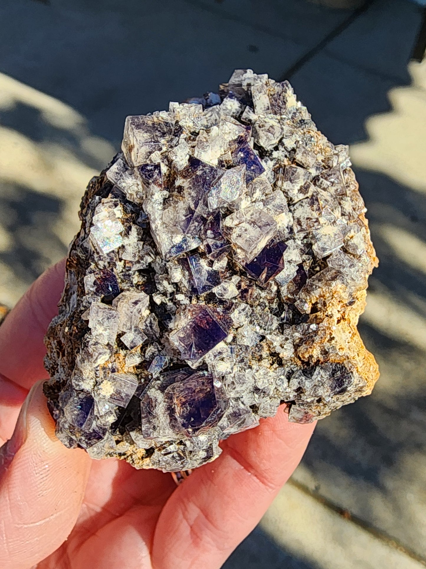 A person holds a raw mineral specimen named Fluorite- Smoky Rabbit Pocket from The Crystalary, mined in the Diana Maria Mine, Frosterley, Weardale, Co. Durham, England. The purple crystals embedded in a rough matrix have a geometric structure that beautifully catches the light for a reflective effect. In the background is Weardale's blurred outdoor setting.