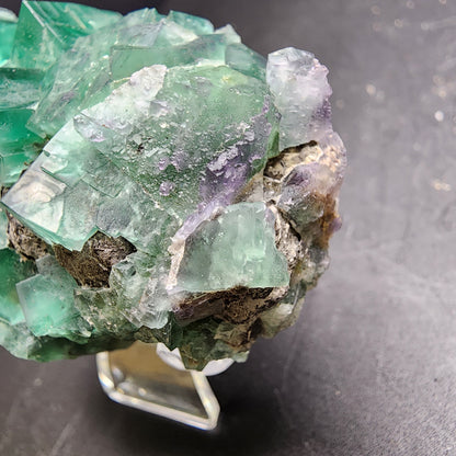 A close-up of a raw Fluorite and Galena crystal from the Greedy Hog pocket at Diana Maria Mine, Co Durham, UK (sku 3067) by The Crystalary sits majestically on a clear stand against a dark background. Its rugged and uneven surface features translucent and opaque areas, showcasing the natural geometric formations that make it truly mesmerizing.