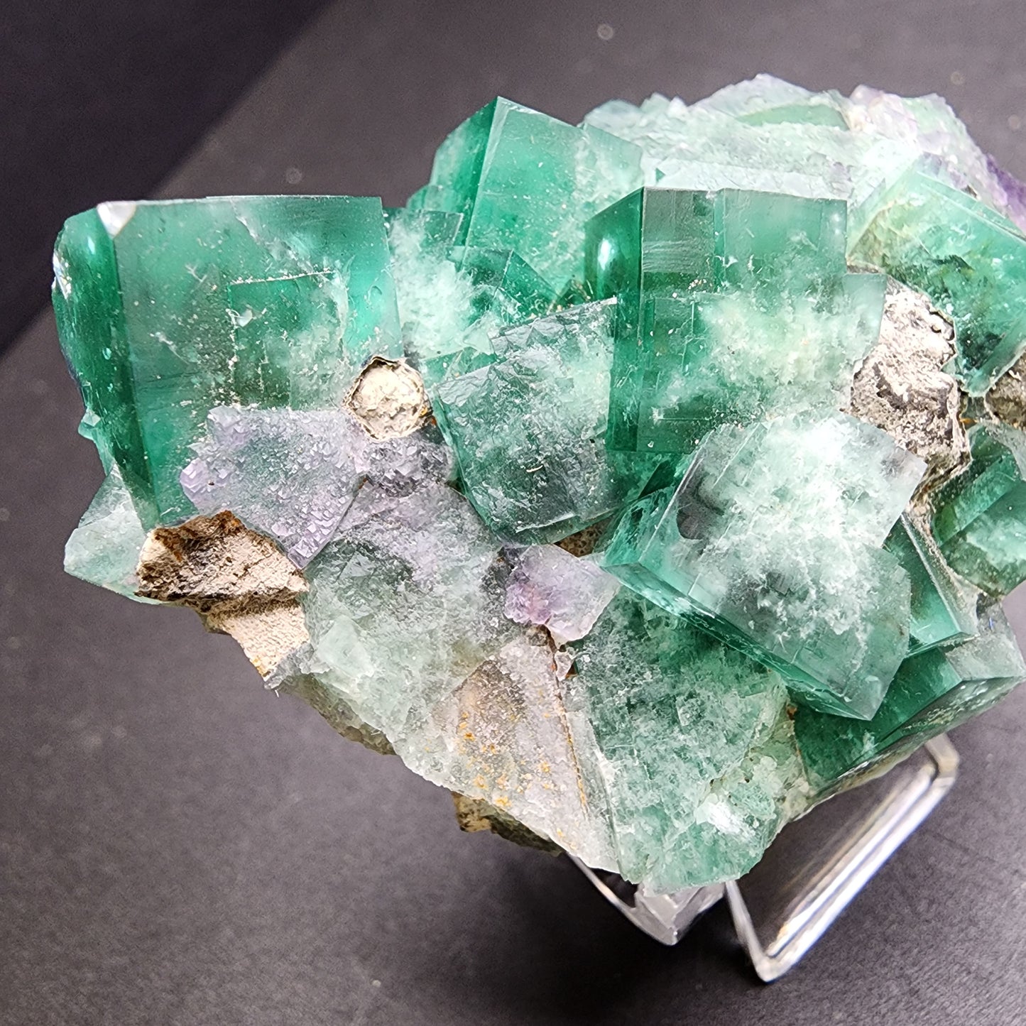 A collection of cube-like green fluorite crystals featuring subtle purple hues from The Crystalary's Fluorite and Galena product (Greedy Hog pocket, Diana Maria Mine, Co Durham, UK) is displayed on a dark surface. These minerals exhibit translucency with natural imperfections that enhance their vibrant color and detailed texture.