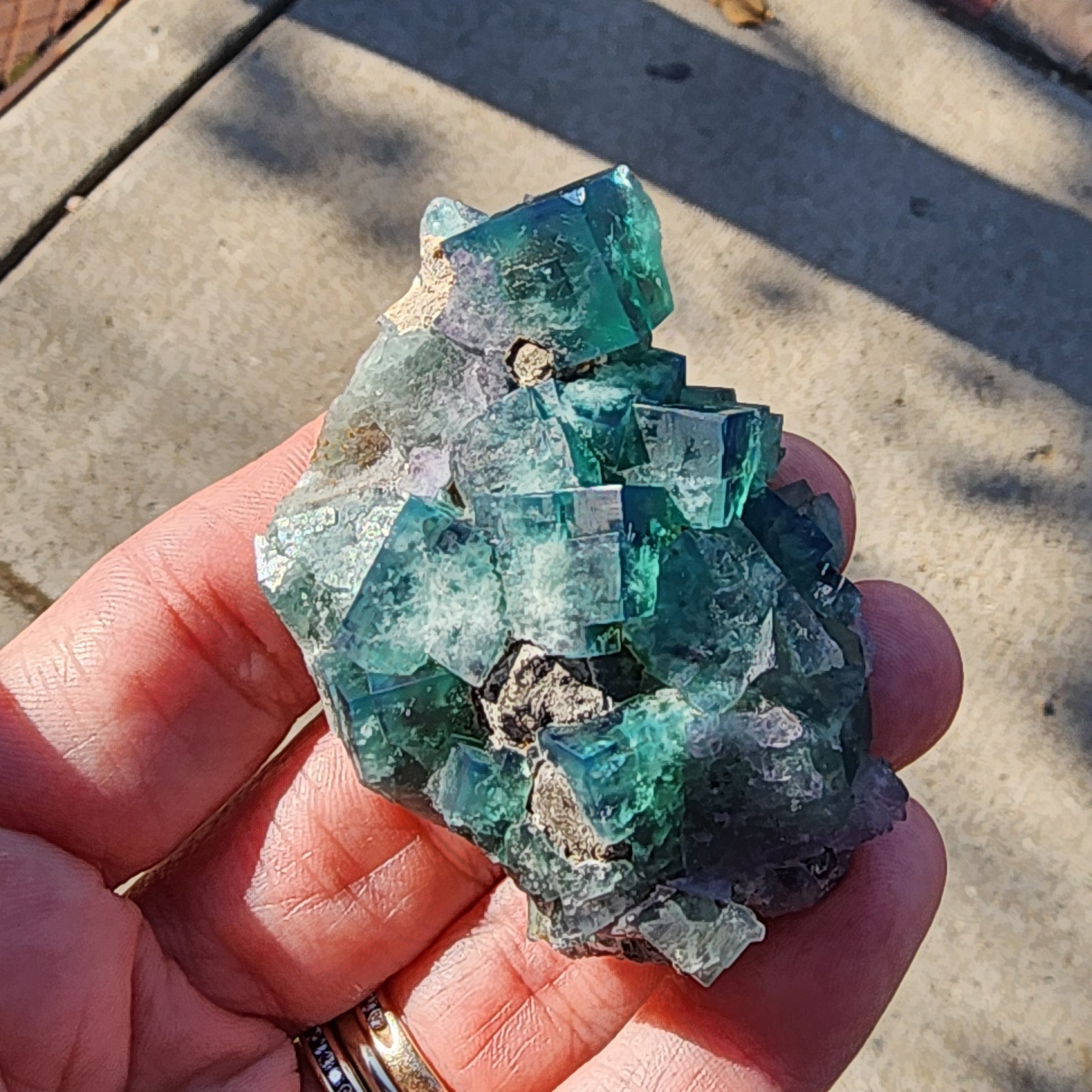 A person holds a mesmerizing cluster of vibrant green-blue fluorite crystals from the Greedy Hog pocket at the Diana Maria Mine, Co Durham, UK. Their rough, jagged texture scintillates under the sunlight. In the background, a blurred outdoor concrete scene unfolds, enhancing the subtle glint of light on the person's ring. The stunning piece is available as Fluorite and Galena from The Crystalary with SKU 3067.