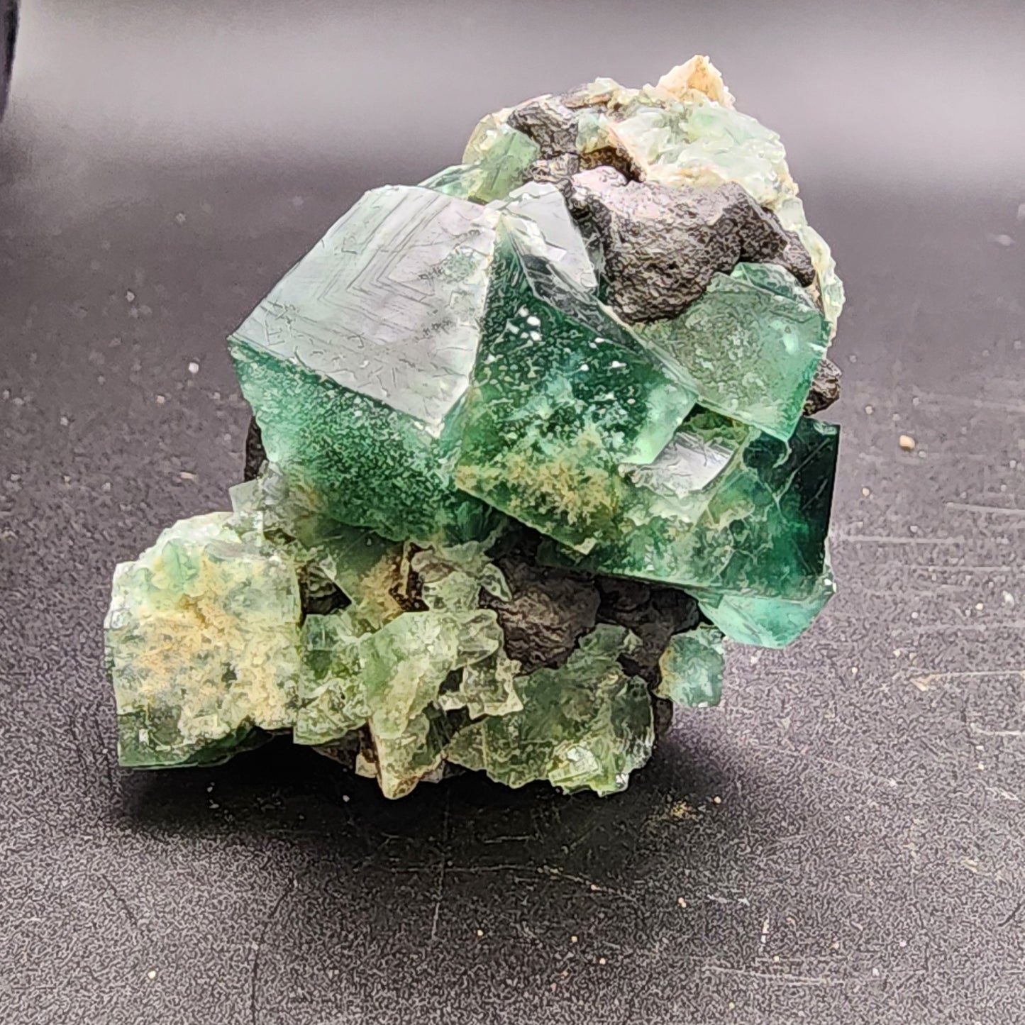 Here's a rephrased version of your sentence using the provided product data:

A close-up view of the raw green cubic crystals of the Fluorite and Galena formation from The Crystalary's Greedy Hog pocket, SKU 3064, at Diana Maria Mine, Co Durham, UK. The crystals exhibit a gentle gradient from light to dark green on their smooth surfaces as they reflect light, all resting atop a dark black Galena matrix base.