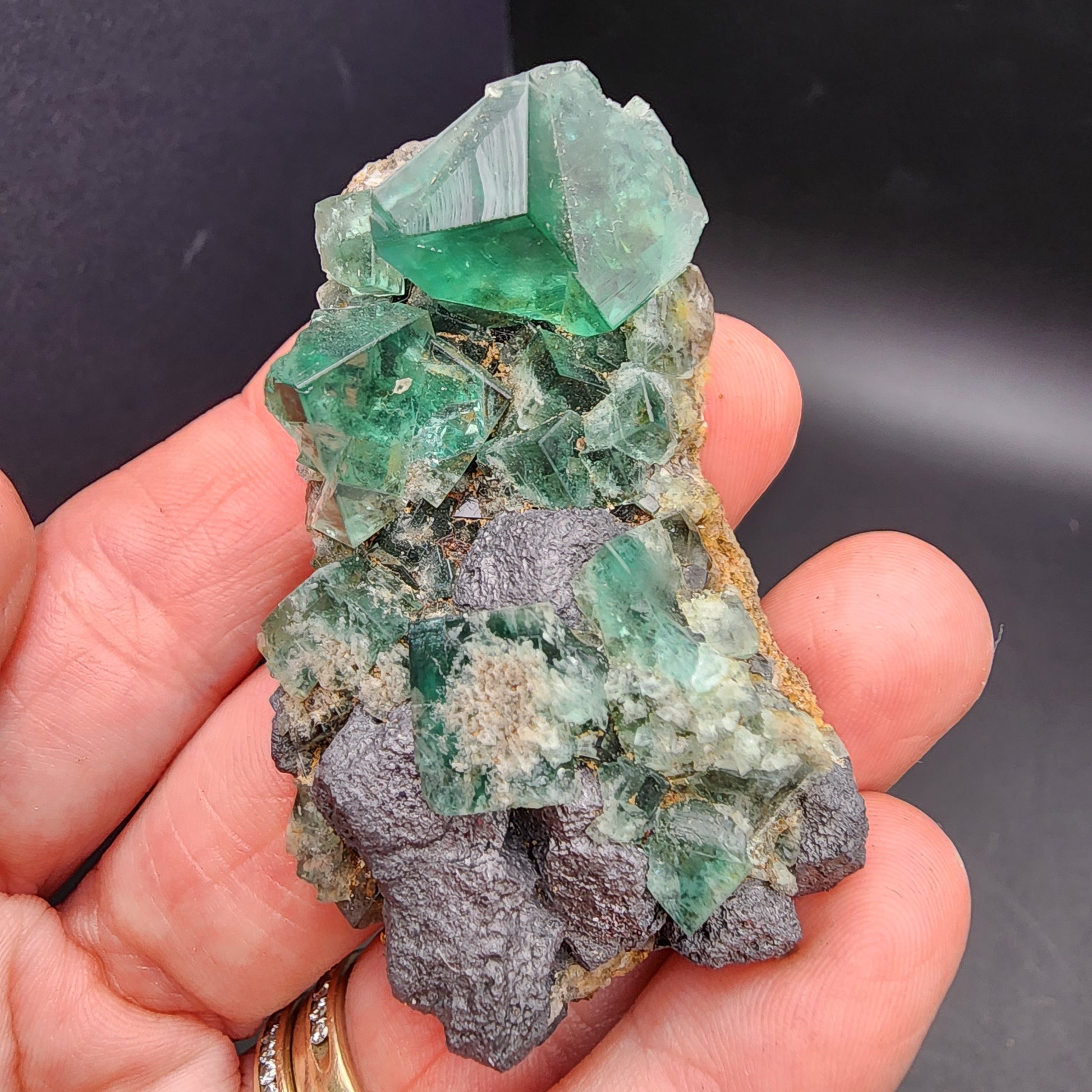 A hand holding a vibrant cluster of green fluorite crystals, sourced from the Greedy Hog Pocket at the Diana Maria Mine in Co Durham, UK. Sold under SKU 3062 by The Crystalary, these semi-transparent crystals exhibit sharp, angular shapes that stand out against their dark brown matrix rock.