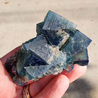 A hand holds a cluster of Fluorite from the Greedy Hog Pocket, sourced from the Diana Maria Mine in Co Durham, UK. The crystals, with sku 3061 and crafted by The Crystalary, shimmer in a blue-green hue with slightly translucent qualities that reveal their rough textures and natural patterns under sunlight.