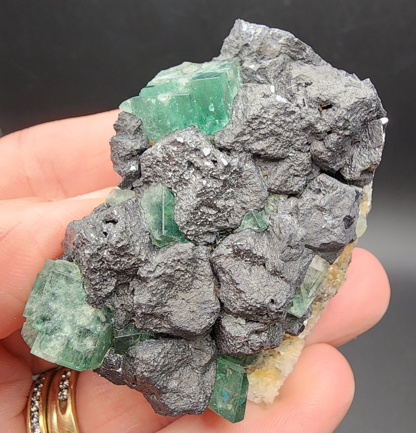 A hand holds a mineral specimen from The Crystalary, labeled "Fluorite and Galena- Northern Lights Pocket, Diana Maria Mine, Co Durham, UK" (sku 3060). It showcases dark, textured rocks interspersed with shiny greenish-blue fluorite crystals. These cubic crystals reflect light in a manner reminiscent of the Northern Lights pocket, offering a striking contrast to the rough surface of the surrounding rocks.