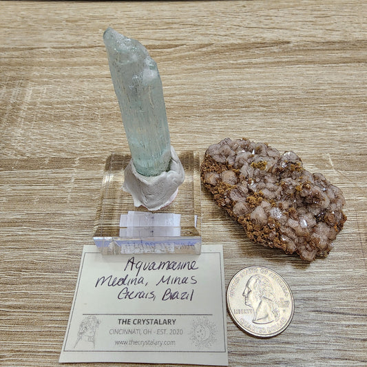 An exquisite aquamarine crystal with a light blue hue, sourced from Medina, Minas Gerais, Brazil, is elegantly displayed on a stand next to a striking brown mimetite cluster. To provide a sense of scale, there is a United States quarter nearby. Accompanied by an informative label from The Crystalary in Cincinnati, OH, this item makes for an extraordinary addition to any collection or giveaway. From the LIVE- VineyardGuy- 11/14/2024 selection by The Crystalary.