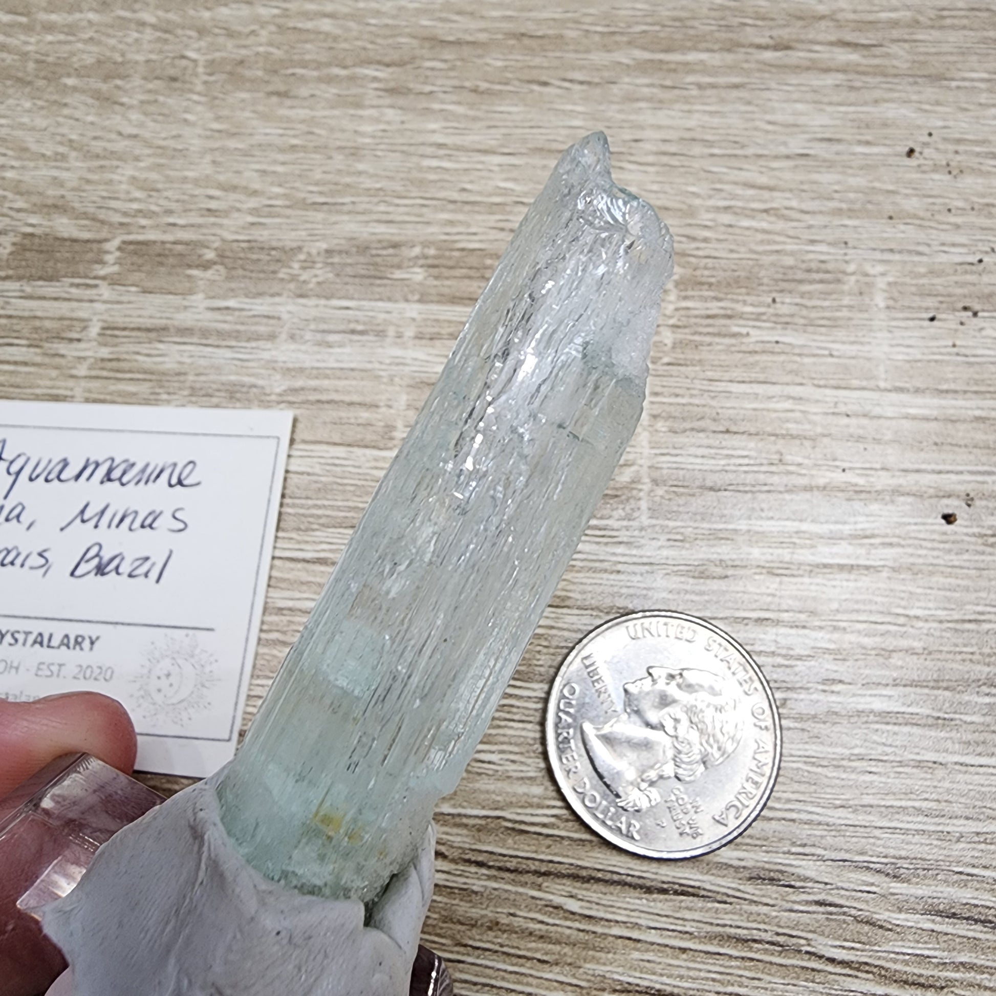 In the photo, a hand displays a transparent aquamarine crystal similar in size to a quarter. The background features a wooden surface alongside the edge of a card with handwritten text, suggesting an enticing giveaway from The Crystalary's LIVE event, VineyardGuy, on November 14, 2024.