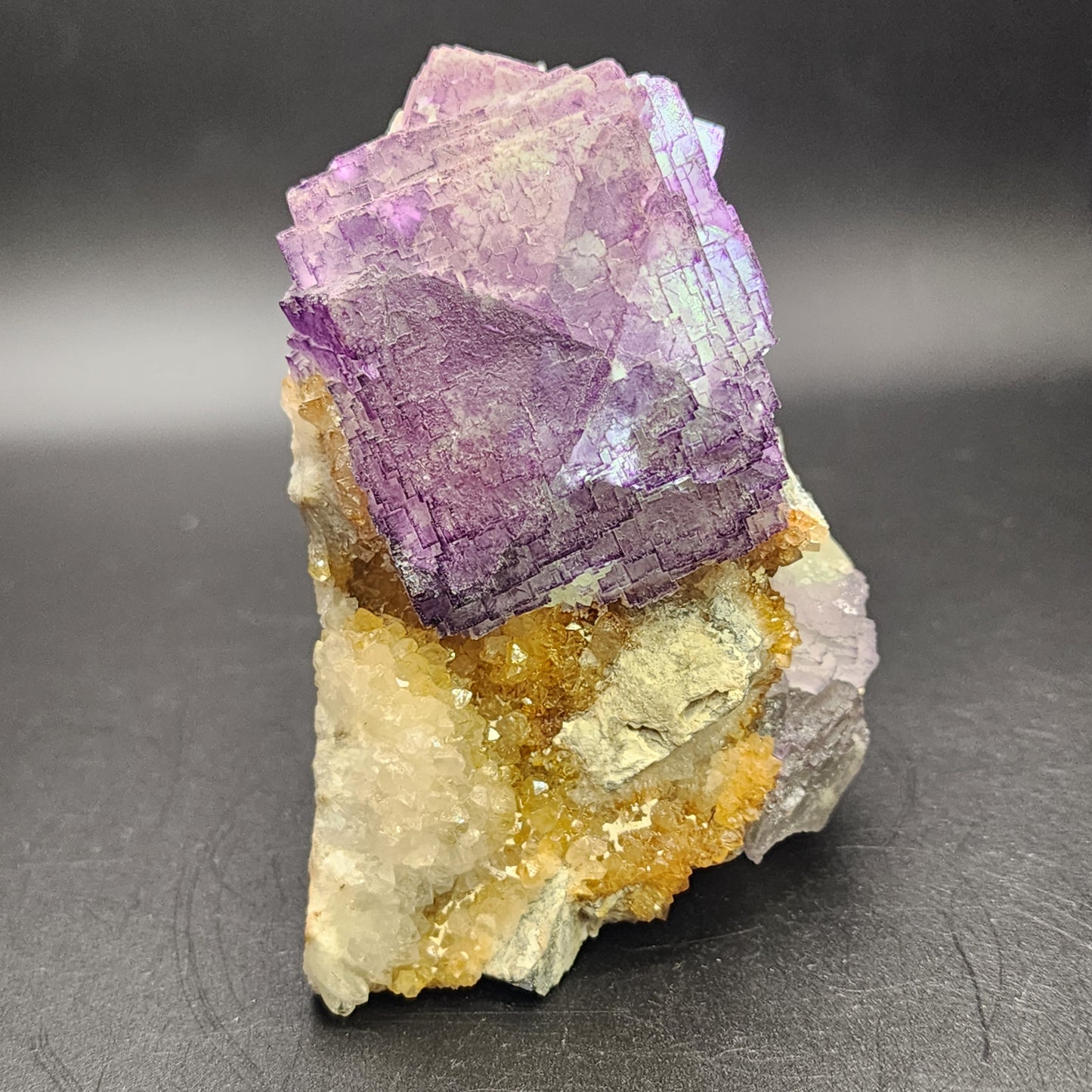 A group of cubic purple crystals, known as Fluorite - Ice Cream Igloo Pocket from Bingham, Hansonburg District in Socorro County, New Mexico, by The Crystalary (sku 3259), sits majestically on a rough rock surface against a dark background. These vibrant purple crystals create a striking contrast with the smaller yellow mineral formations below, evoking the rich mineralogical history of New Mexico.