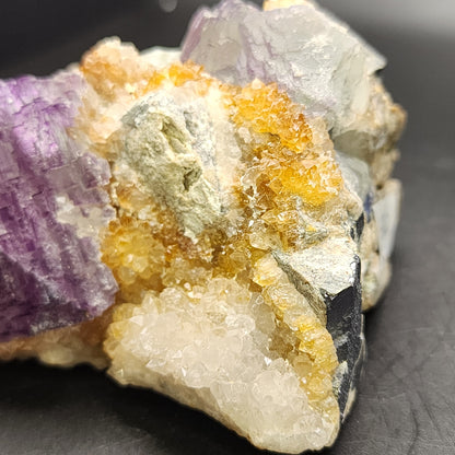 Featured in the image is a close-up of "Fluorite- Ice Cream Igloo Pocket" from The Crystalary, showcasing a US specimen from Bingham, Hansonburg District, Socorro County, New Mexico (sku 3259). This specimen features an intriguing mix of purple, yellow, and white crystal formations. Its rough surface texture reveals various layers and shades set against a dark background.