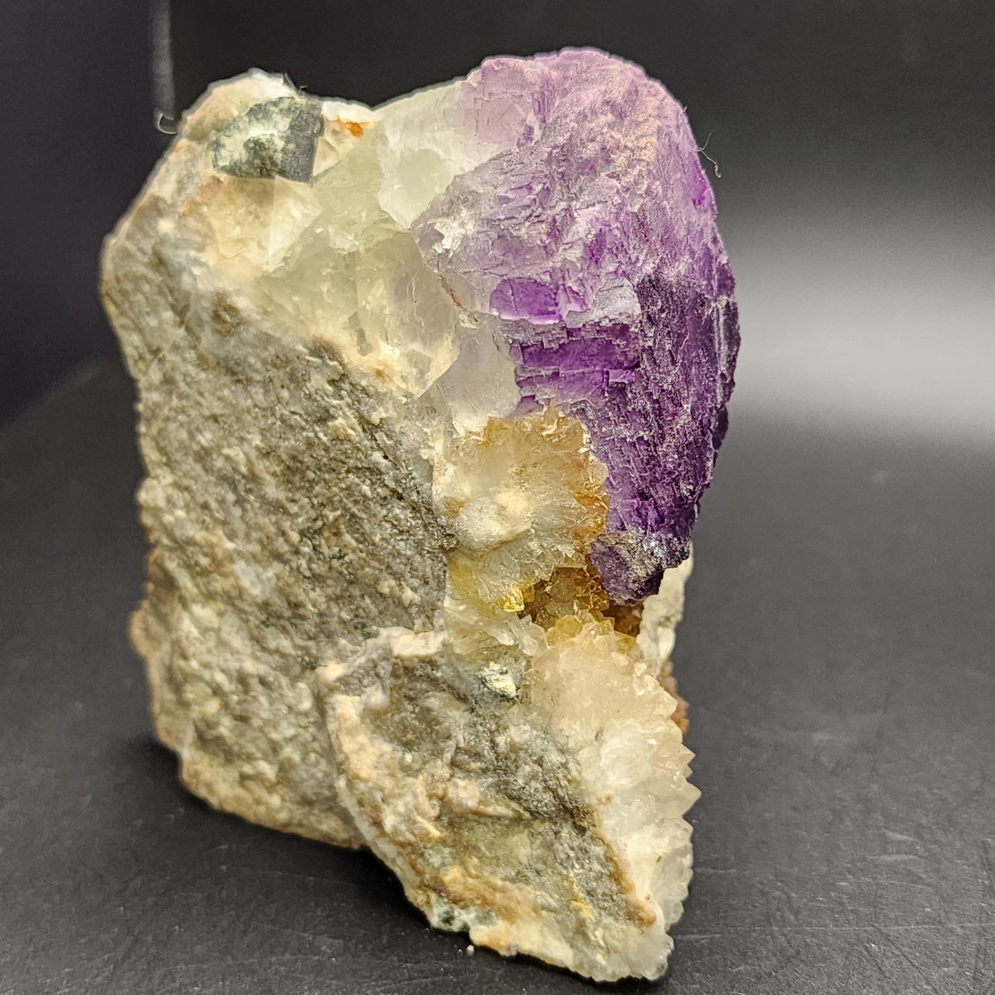 An unrefined mineral piece features a vibrant purple amethyst embedded in a light gray rock, reminiscent of New Mexico's geological character. It is set against a dark background and flaunts the clear, angular structure characteristic of The Crystalary's "Fluorite- Ice Cream Igloo Pocket" from the Bingham, Hansonburg District in Socorro County, New Mexico (SKU 3259).