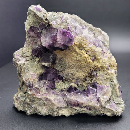 A substantial, coarse rock featuring aggregates of purple amethyst crystals against a textured gray and sandy base, evocative of the remarkable specimens typically available at The Crystalary, set on a plain dark background.