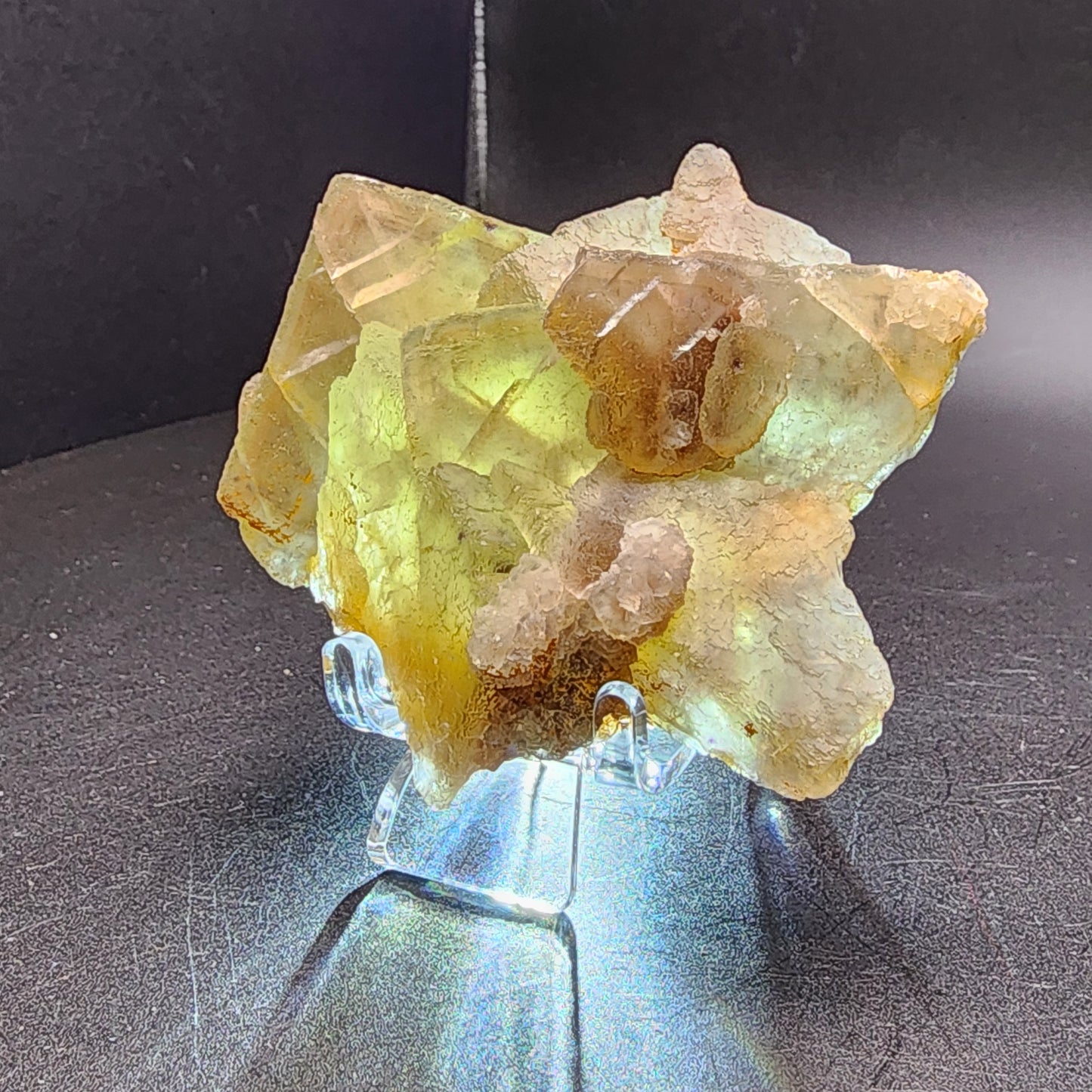 An arrangement of translucent yellow and brown phantom fluorite crystals from the Heißer Stein Mine, showcased on a small stand, glistens with a glossy finish. Illuminated from below, these intricate formations from the SKU 3080 collection by The Crystalary are beautifully highlighted against a dark background.