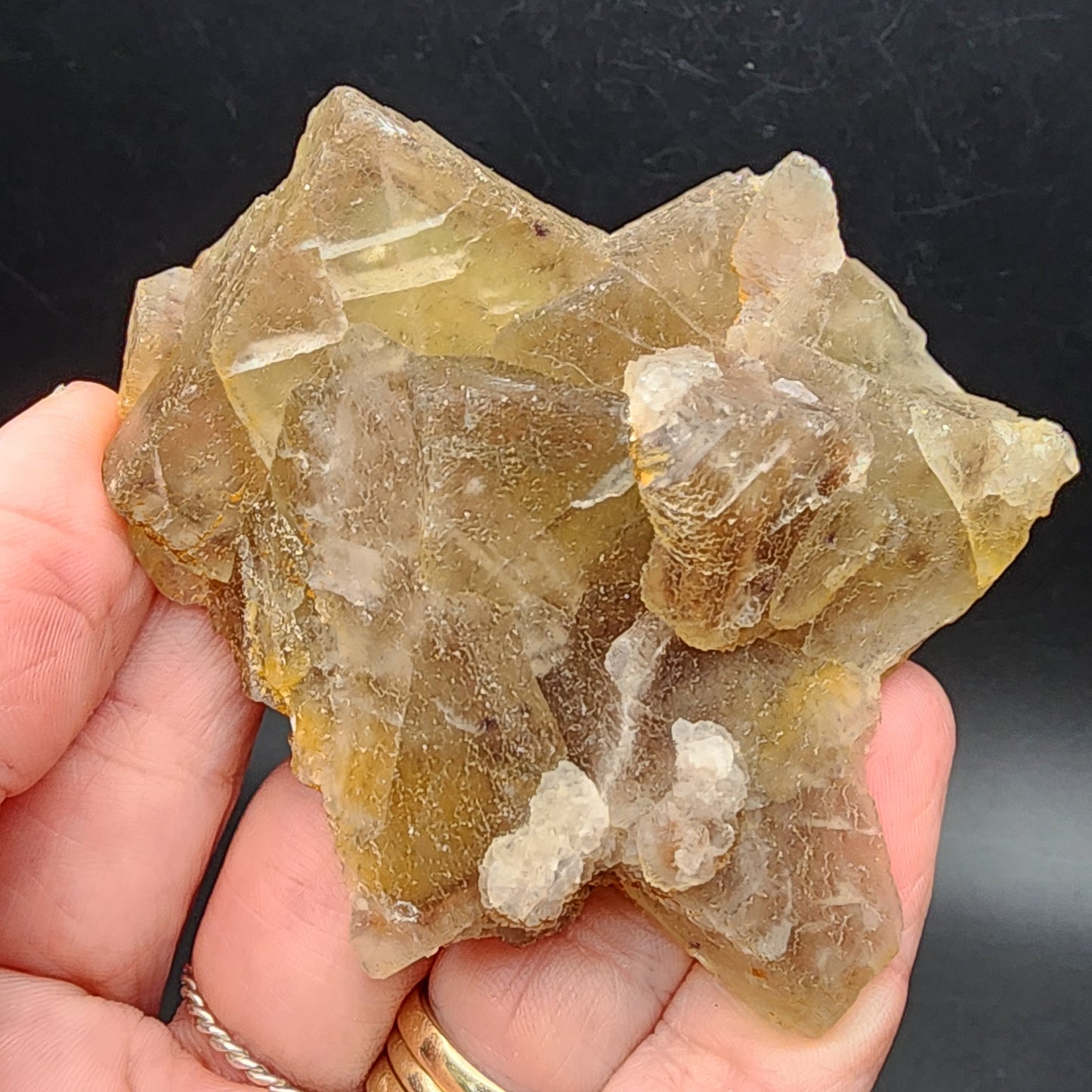 A hand displays a cluster of translucent, brownish phantom fluorite crystals with distinct geometric shapes and rough edges from The Crystalary's collection, specifically the Fluorite- Heißer Stein Mine, Stulln, Schwandorf District, Upper Palatinate, Bavaria, Germany (SKU 3080), set against a dark background.