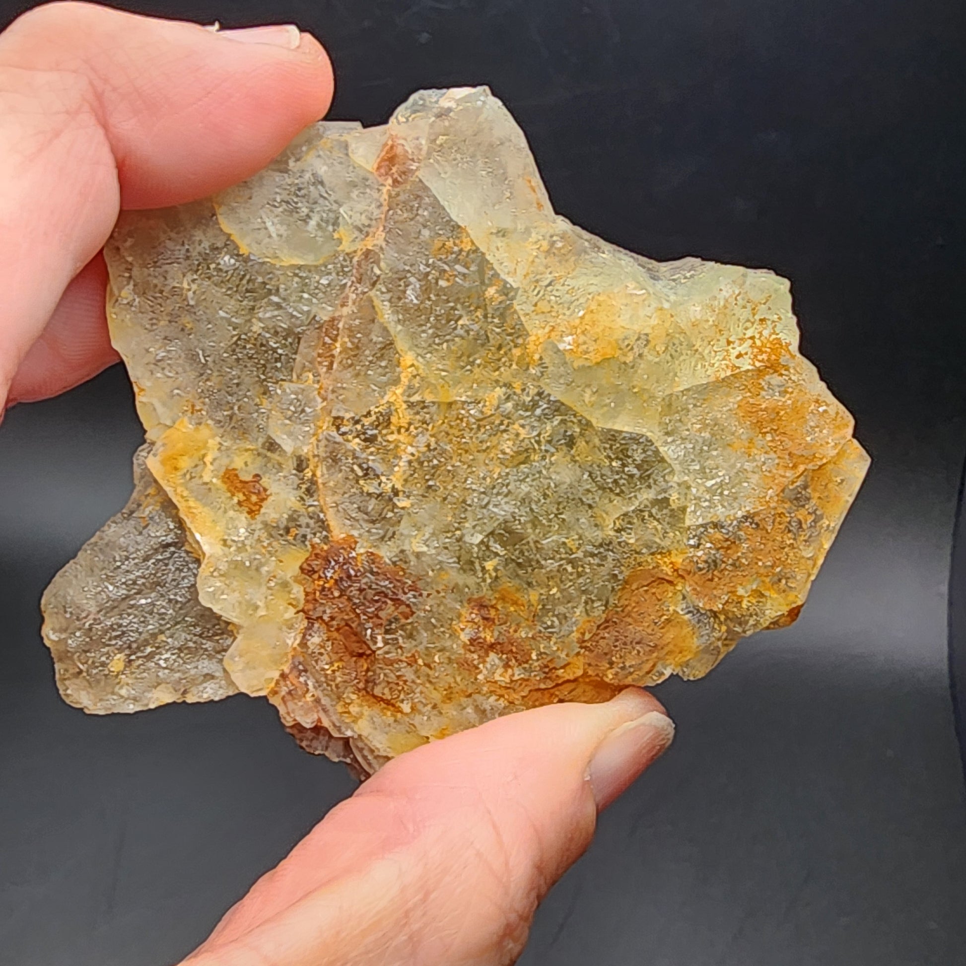 A hand holds a large, irregularly shaped crystal with a rough texture, resembling the rare Fluorite from Heißer Stein Mine. This specimen shows shades of gray, yellow, and orange against a dark background, reminiscent of treasures from The Crystalary (sku 3080).