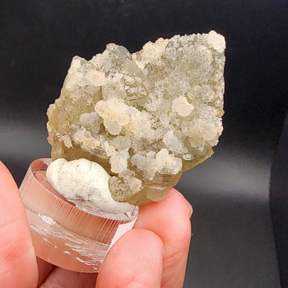 Close-up of a person holding "Fluorite/Quartz- Hermine Mine," product sku 3081, from The Crystalary, sourced from Lissenthan, Nabburg, Schwandorf District in Upper Palatinate, Bavaria, Germany. It is displayed on a clear stand against a dark background. This translucent crystalline specimen features a rough texture with small clustered formations.