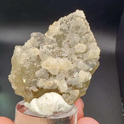 A hand holds a rough, translucent Fluorite/Quartz crystal, highlighting its textured and shimmering appearance as it sparkles on a clear stand against a dark background. Sourced from the Hermine Mine in Lissenthan, Nabburg, Schwandorf District, Upper Palatinate, Bavaria, Germany, this stunning specimen is part of The Crystalary’s collection with SKU 3081.