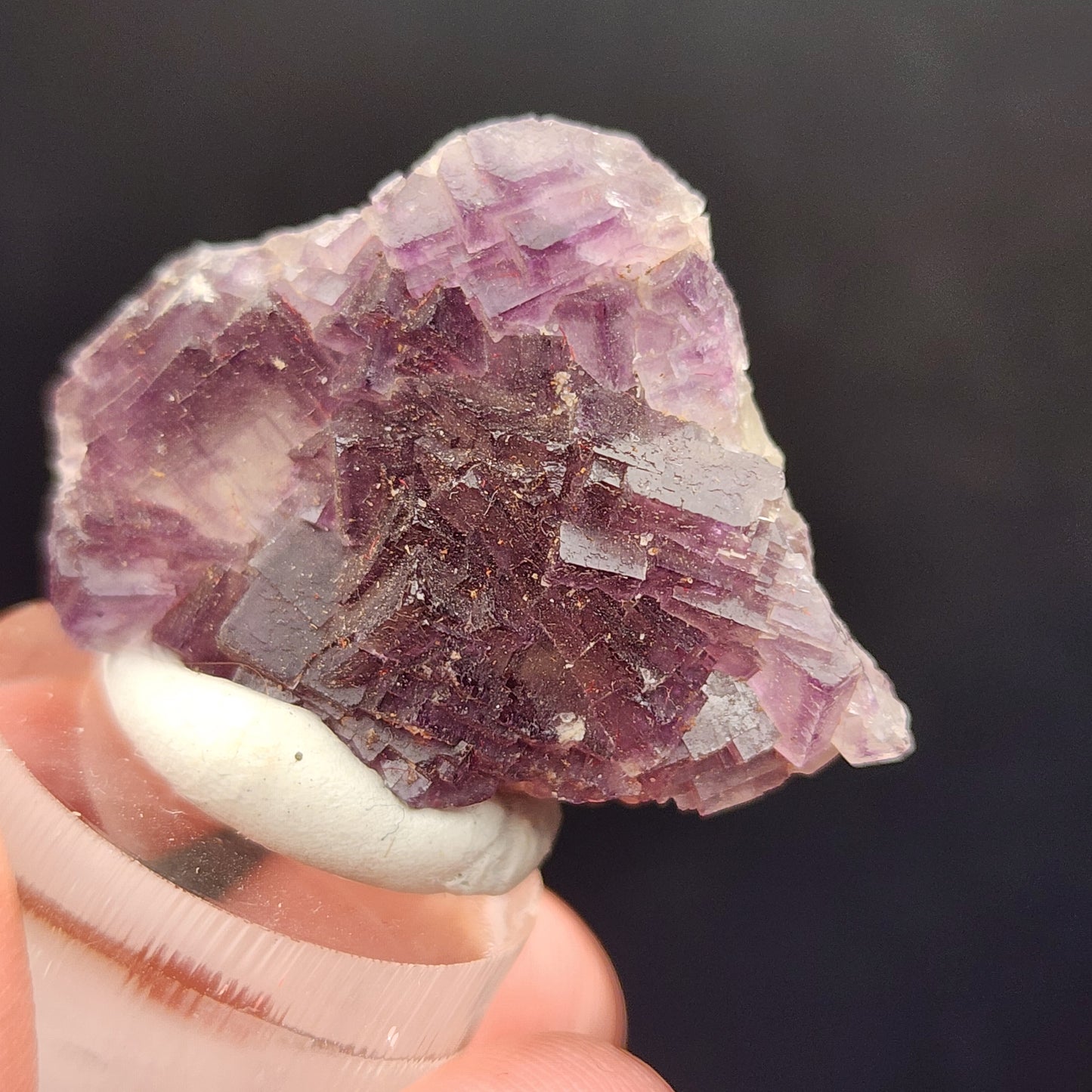 A close-up image captures a raw purple fluorite miniature with cubic formations held in someone's hand against a dark background. This exquisite crystal from The Crystalary, sourced from the Hermine Mine in Lissenthan, Nabburg, Schwandorf District, Upper Palatinate, Bavaria, Germany (sku 3082), showcases varying shades of purple alongside some white areas.