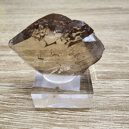 A large, transparent brown smokey quartz crystal from The Crystalary's LIVE- HappytobeMe1016- 12/12/2024 collection, reminiscent of a Tiefen Glacier Gwindel, with jagged edges sits on a clear square stand against a light wooden surface.