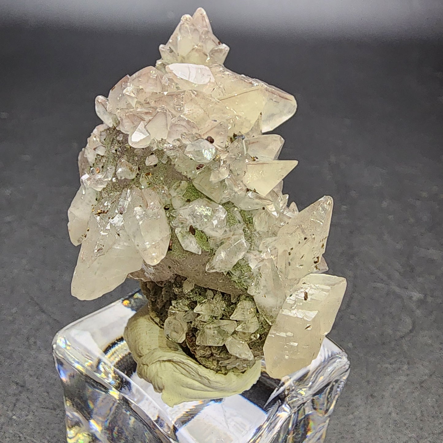 A cluster of clear and light pink crystals resembling amethyst from The Crystalary is displayed on a stand. These intricately jagged quartz and calcite formations are sourced from Tonglushan Mine, Daye Co., China, and contrast beautifully against a plain dark surface (SKU: 3100).