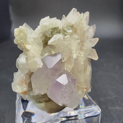 The Crystalary's Quartz v. Amethyst, Calcite from Tonglushan Mine (sku 3101) is displayed on a reflective square base against a dark background. The amethyst and purple crystals, with sharp edges and varying sizes, shimmer subtle greenish tinges.