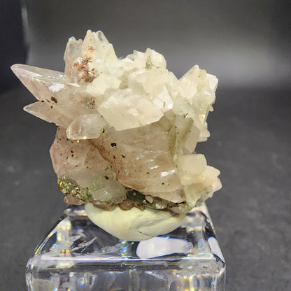 The Crystalary's Quartz v. Amethyst with Calcite from Tonglushan Mine (sku 3101) is elegantly showcased on a clear glass stand, set against a dark background, featuring clear quartz crystals with brown and green inclusions.