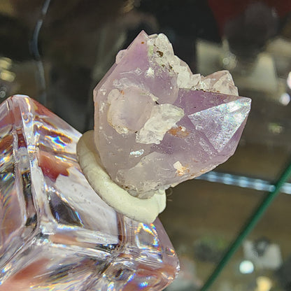 The Crystalary's Quartz v. Amethyst (sku 3102) from Tonglushan Mine is artfully displayed on a glass bottle, secured with clay. It showcases purple hues with white inclusions, and its facets are illuminated alongside calcite accents for visual appeal.