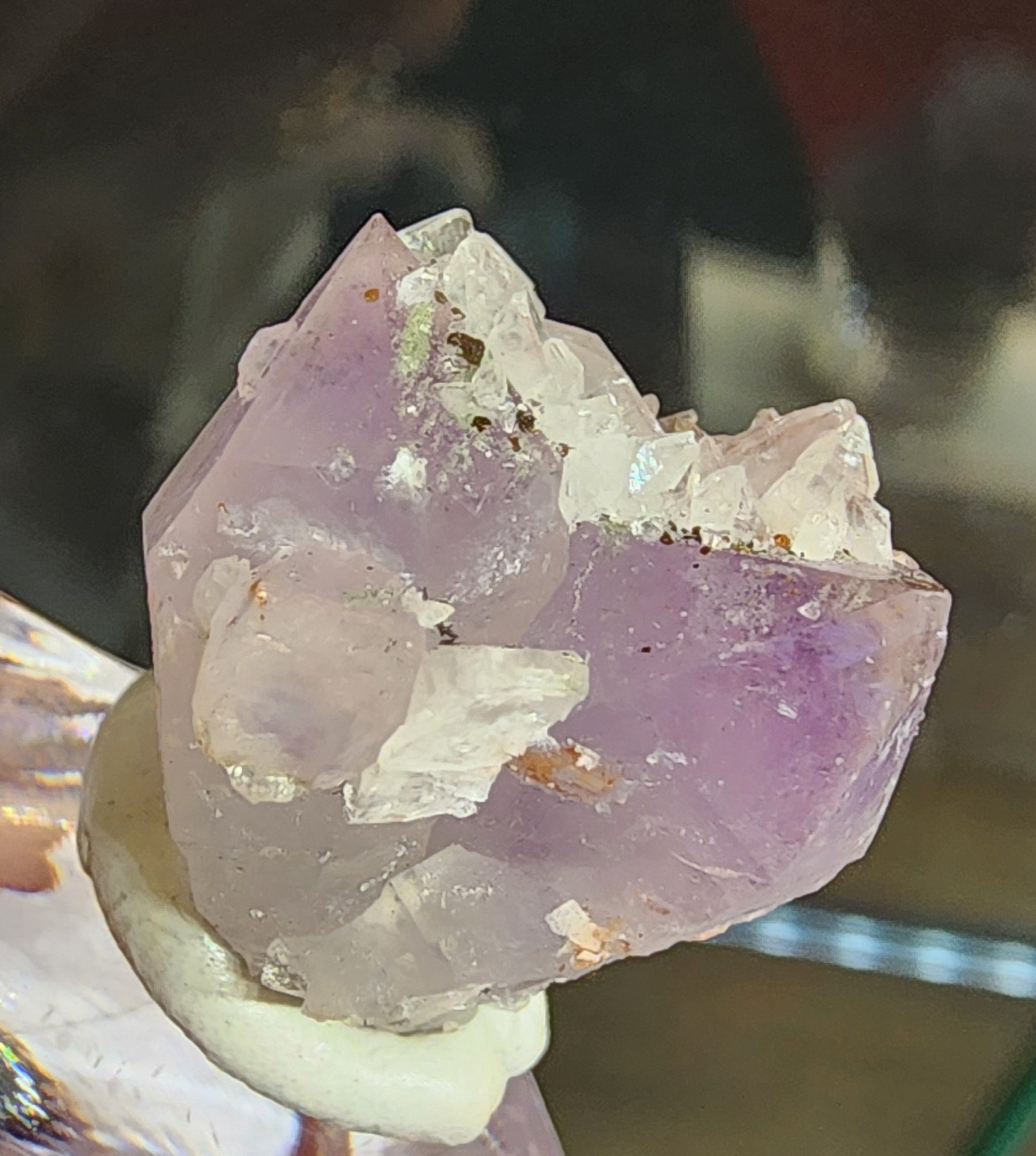 The Crystalary's product, Quartz v. Amethyst, Calcite (sku 3102), exhibits a purple and white amethyst crystal cluster with jagged edges on a white base from Tonglushan Mine. The lighting enhances its transparency and texture against a blurred background.