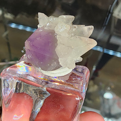 A hand holds The Crystalary's Quartz v. Amethyst, Calcite vial (sku 3103) featuring amethyst and white crystals from the Tonglushan Mine, China. The light casts a rainbow on the glass, highlighting the crystals' intricate surfaces reminiscent of hidden treasures.