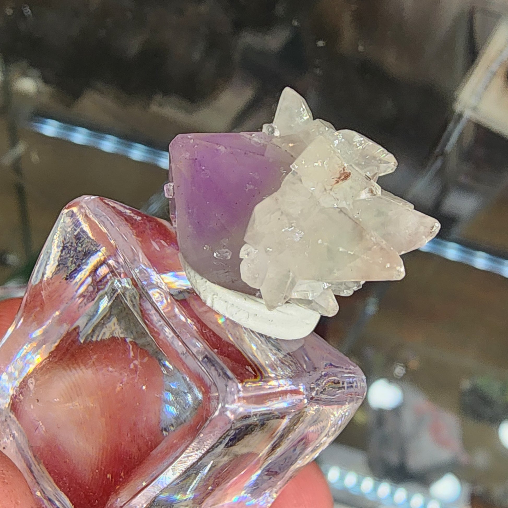 Someone holds a small, clear glass bottle by The Crystalary (sku 3103) featuring a crystal cluster of Quartz and Amethyst with purple and clear geometric shapes attached to the cap. The blurred background highlights the intricate details of these stunning formations from Tonglushan Mine, China.