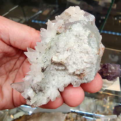 A person holds a cluster of clear quartz crystals from The Crystalary, featuring pointed, translucent formations. Violet hints of amethyst add to the rough texture with white and greenish areas. The display case in the background contains calcites from the renowned Tonglushan Mine (sku 3105).