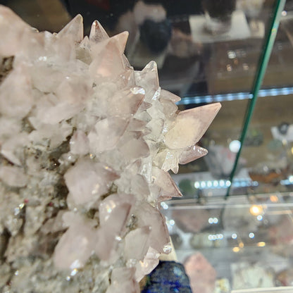 A cluster of pinkish quartz crystals from The Crystalary, SKU 3105, is showcased in front of a glass cabinet. Amethyst and calcite specimens from the Tonglushan Mine enrich the display, highlighting their sharp formations.