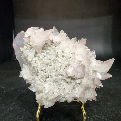 A metallic stand supports a cluster of clear quartz crystals against a dark backdrop. The sharp, pointed formations are semi-transparent and evoke the Quartz v. Amethyst, Calcite from Tonglushan Mine by The Crystalary (sku 3105), showcasing varied angles and surfaces.