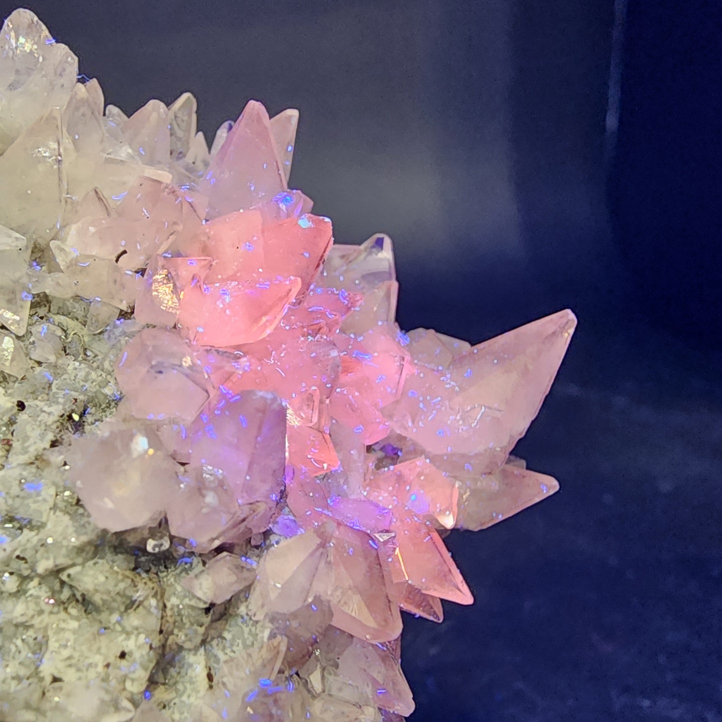 This Quartz v. Amethyst and Calcite cluster from The Crystalary (sku 3105) features pointed pink formations with faint iridescent blue specks, set against a textured rock base. It creates a vibrant contrast reminiscent of minerals found in the Tonglushan Mine, Daye Co., China.