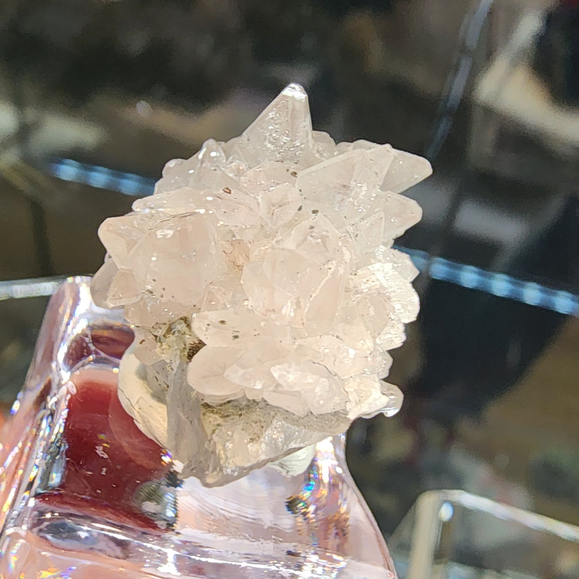 The Crystalary's Quartz v. Amethyst with Calcite (SKU 3106) from Tonglushan Mine displays translucent white crystals with sharply pointed formations, intricately arranged to reflect light, reminiscent of miniature sculptures on a shiny surface with a blurred background.