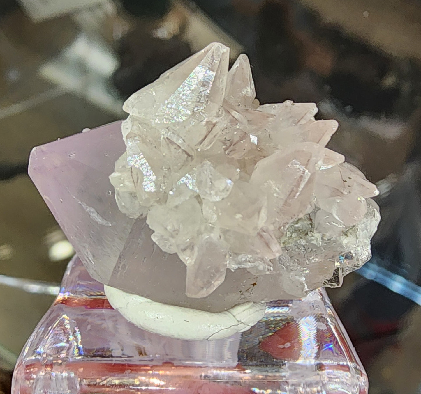 A close-up of "Quartz v. Amethyst, Calcite" from The Crystalary showcases translucent, jagged formations with a pale pink hue on a reflective surface. Resembling a treasure from the Tonglushan Mine, it is elegantly mounted on a small white base. SKU 3106.