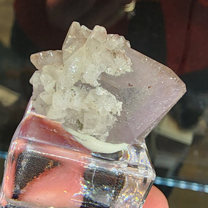 A hand holds a transparent display box containing a large, pale purple amethyst cluster reminiscent of Tonglushan Mine formations. This Quartz v. Amethyst, Calcite piece from The Crystalary (sku 3107) rests atop a cubic base, set against a blurred background.