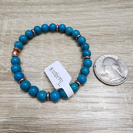 A turquoise beaded bracelet, showcasing sleeping beauty turquoise and copper accents, is artfully arranged on a wooden surface. A white tag reads "LIVE- Winzalite- 1/2/2025" from The Crystalary. For size reference, a quarter is positioned next to the bracelet.