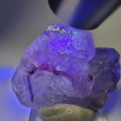 A close-up of The Crystalary's Fluorite- Huallanca, Ancash, Peru (sku 804) presents a cluster of translucent crystals. When lit, they display vibrant colors and intricate structures, elegantly resting on a small stone.