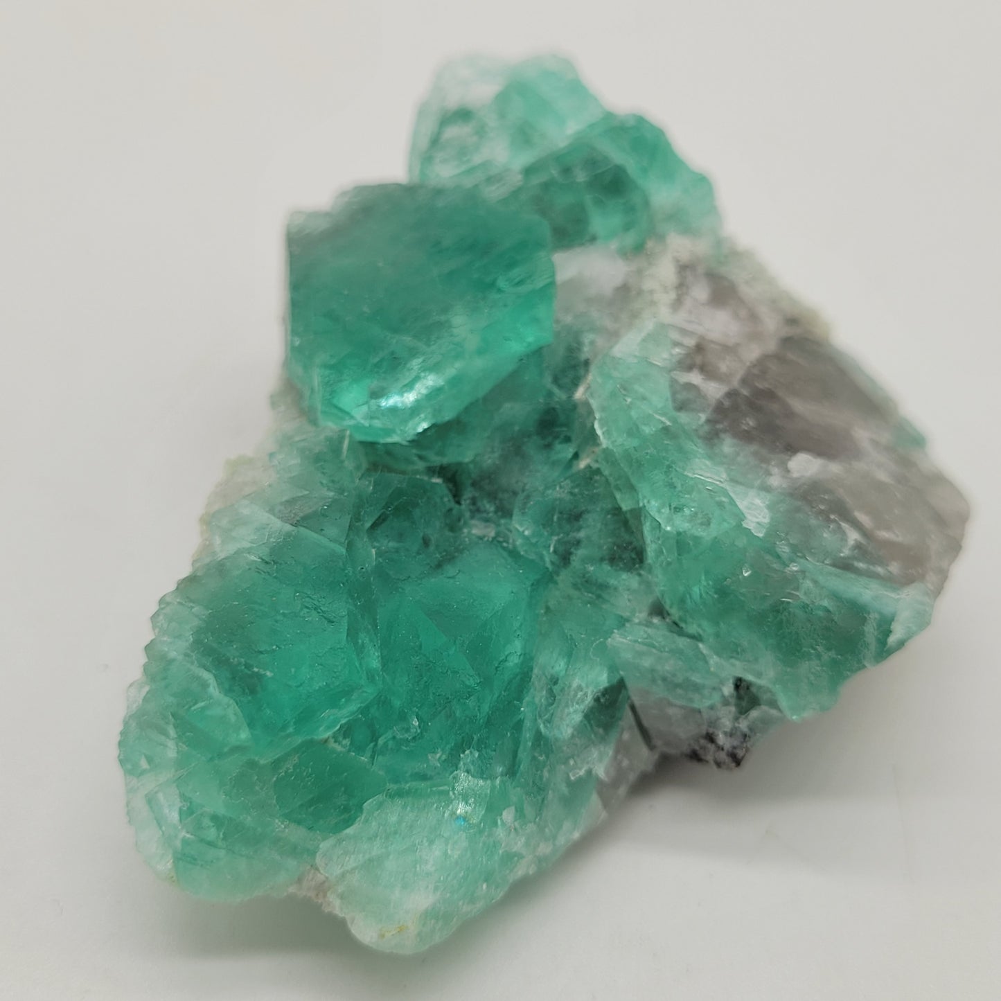 The Crystalary's Fluorite-El Papiol from Sant Cugat del Valles, Barcelona, Catalunya (SKU 805) features translucent raw minty green crystals with vivid color on a light background. The irregular shapes and natural textures enhance the unique beauty of this fluorite specimen.