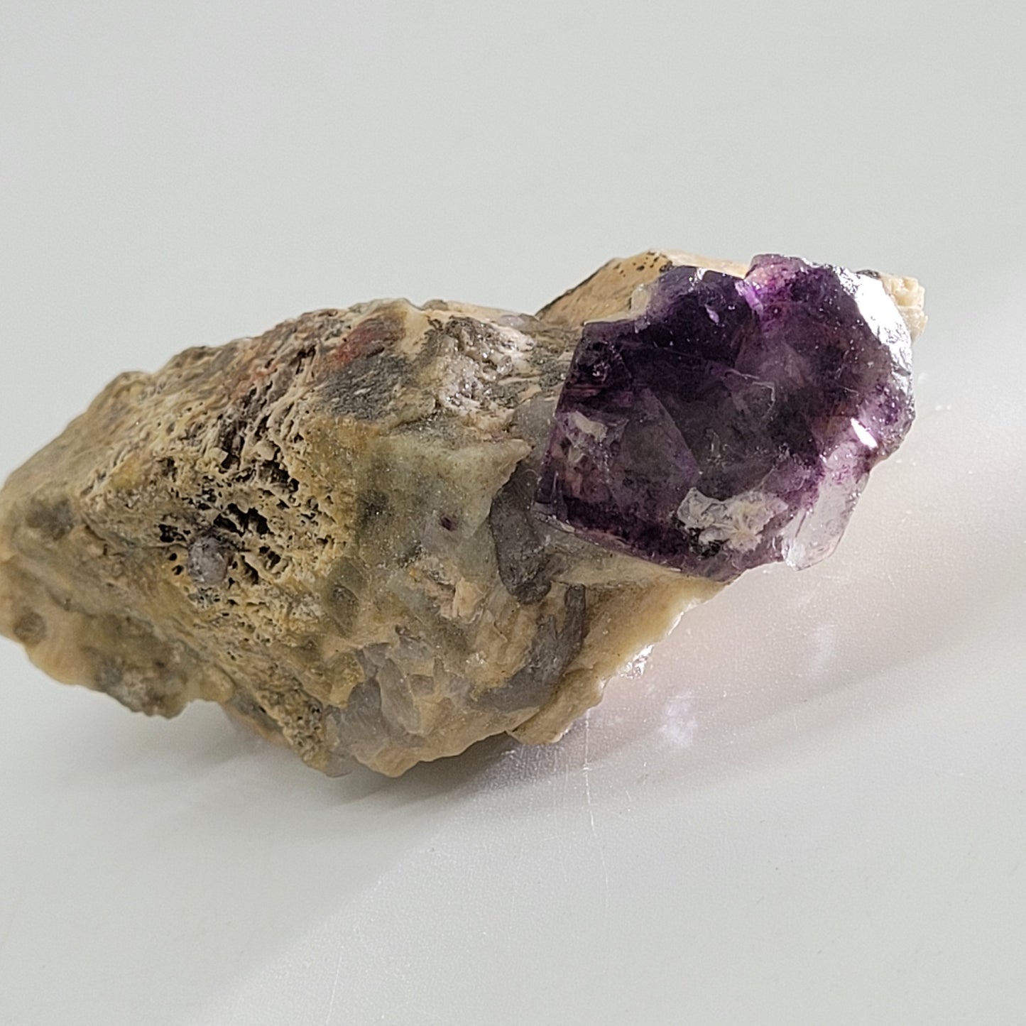 A rough stone, "Fluorite, Feldspar- Minas Gerais, Brazil" SKU 807 from The Crystalary, showcases a prominent purple fluorite crystal with vibrant hues and faceted edges on a light surface.