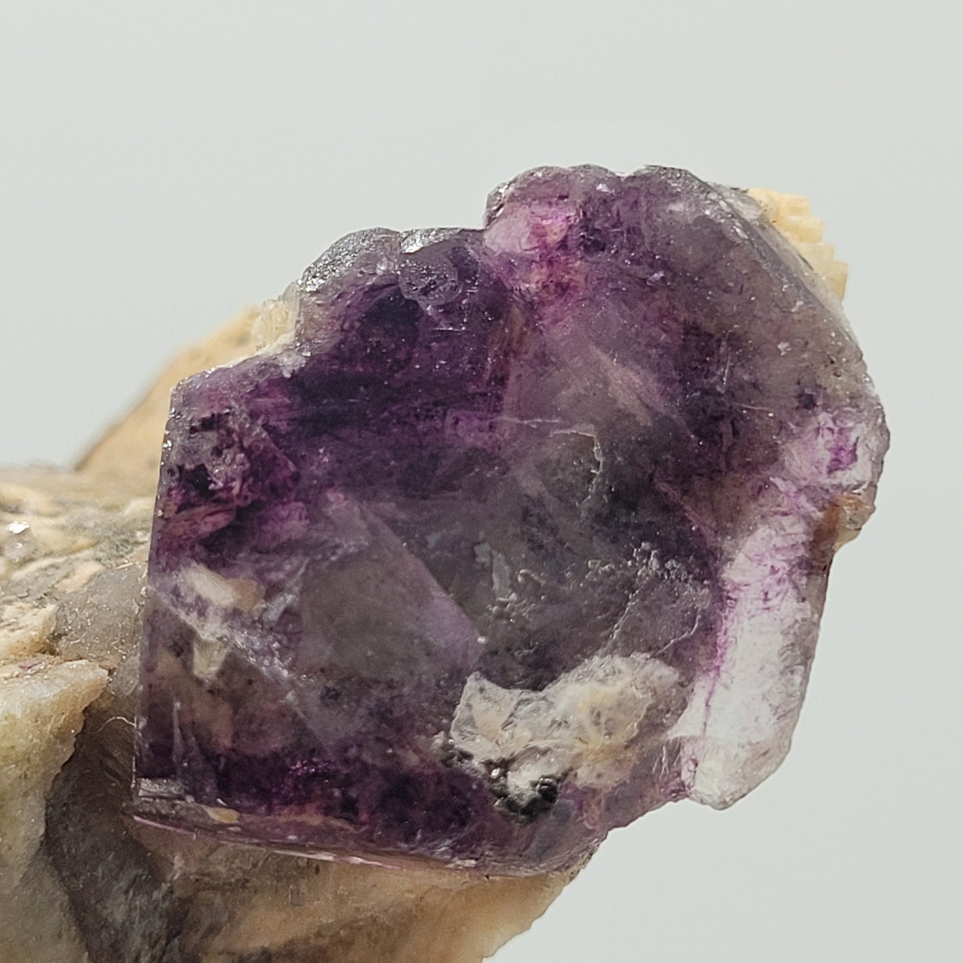 This Fluorite from The Crystalary, sku 807, features a rough, semi-transparent purple fluorite crystal with jagged edges. Embedded in rock from Minas Gerais, Brazil, it displays deep purple and lavender hues like a stained glass window, alongside earthy tones.