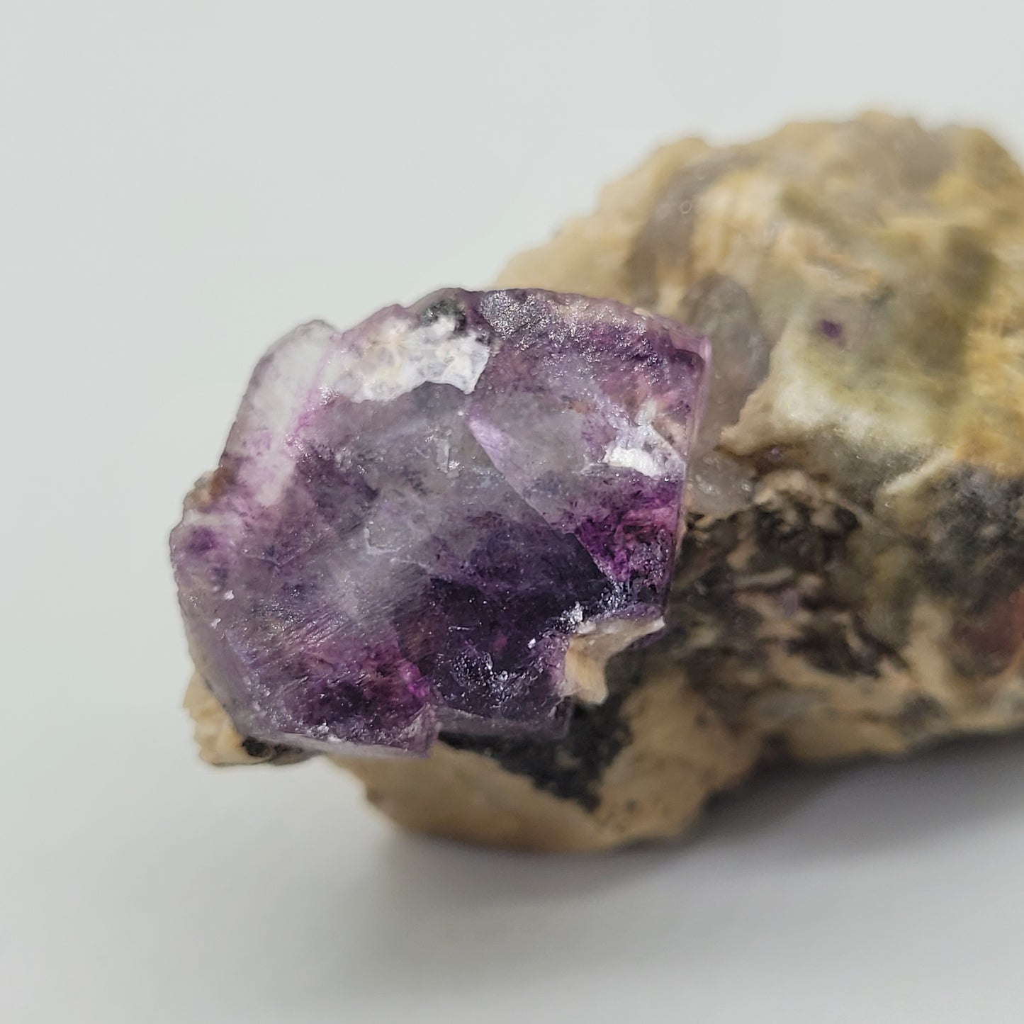 The Crystalary's Fluorite, Feldspar from Minas Gerais, Brazil (sku 807) features a vibrant raw purple fluorite crystal on a beige stone base. The translucent cubic structure displays lilac and deep purple shades with a stained-glass-like beauty and reflective surface.