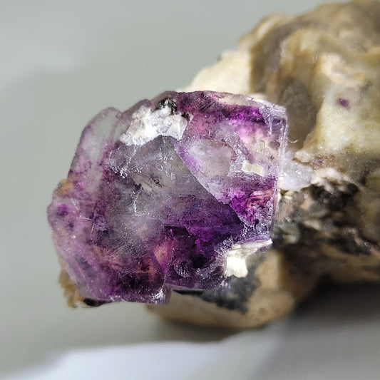 Close-up of "Fluorite, Feldspar- Minas Gerais, Brazil" by The Crystalary, featuring a raw translucent purple mineral crystal in rough beige rock. Natural facets display white and deep violet tones against a softly blurred background. SKU: 807.