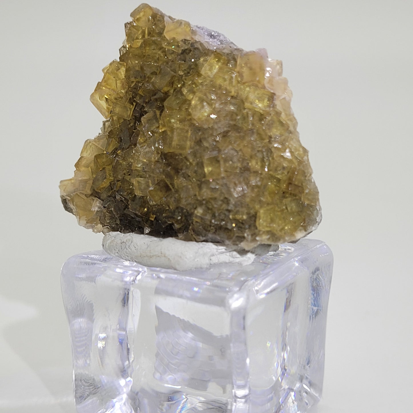 A close-up of The Crystalary's miniature honey yellow fluorite crystal cluster, available as sku 809 from the renowned Moscana Mine in Corvera de Astruias, Spain, is elegantly displayed on a transparent cube stand. Its angular formations and rough texture shine against a plain light background.