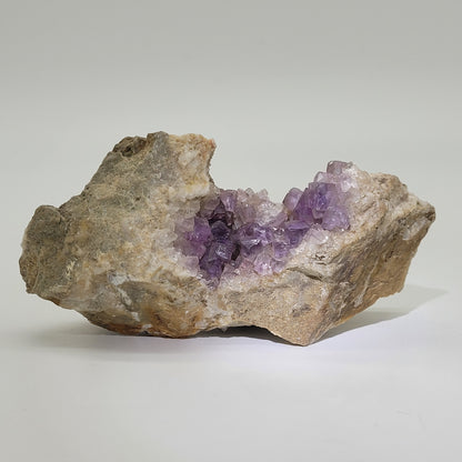 Displayed against a plain backdrop is a natural rock specimen from The Crystalary, featuring a cavity filled with sparkling purple amethyst-like crystals reminiscent of the ones found in Asturias, Spain, SKU 810.