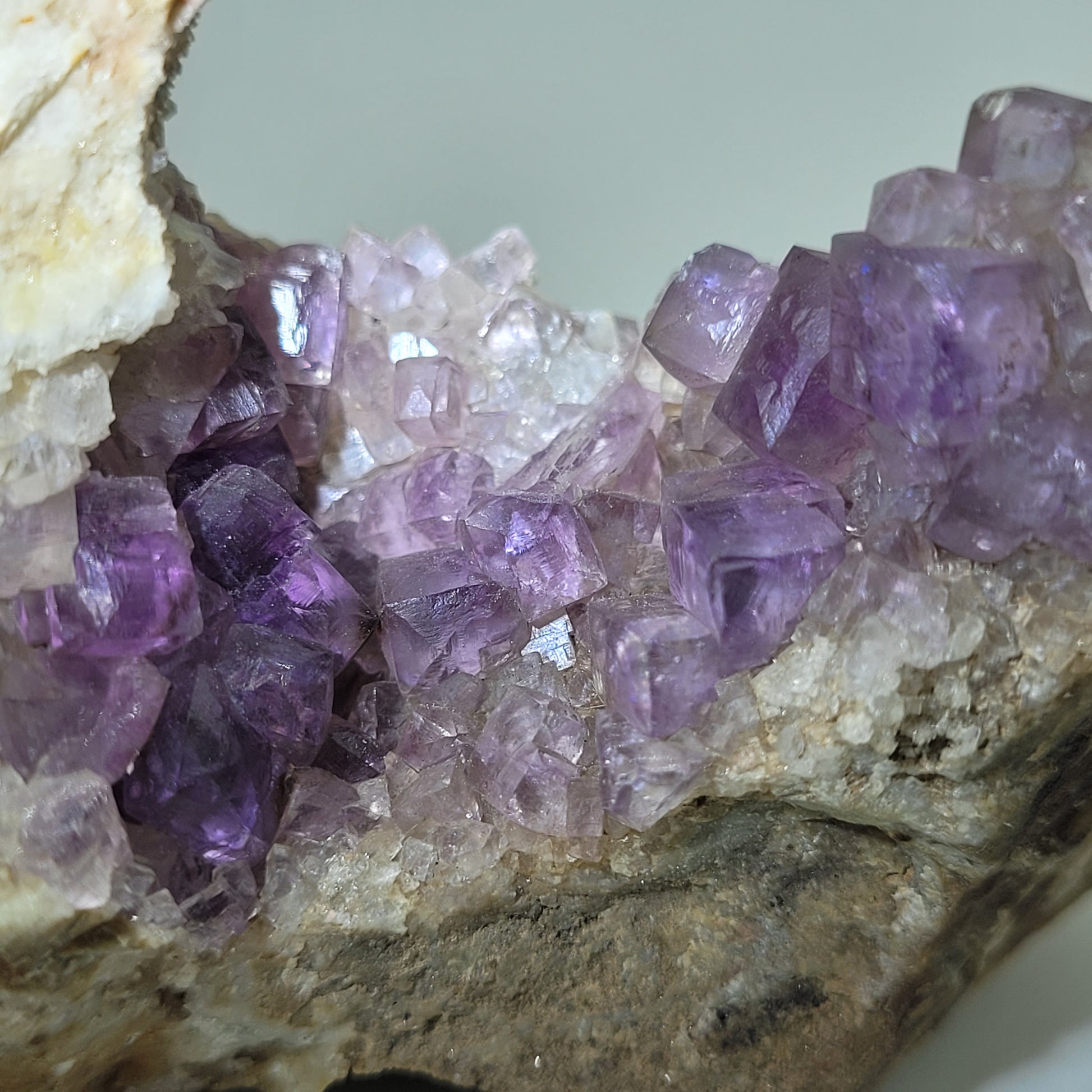 Close-up of The Crystalary's purple cubic fluorite crystals from the Jaimina Mine in Spain (SKU 810), set in light-colored rock. These crystals range from light to deep purple, offering a translucent sparkle against the textured surface.