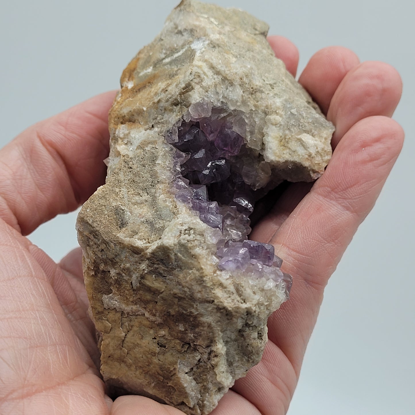 A hand holds a rough specimen rock with Fluorite crystals from the Jaimina Mine, Asturias, Spain. The Crystalary's piece showcases shiny facets contrasting its rough beige exterior, reminiscent of treasures found in Spanish localities. SKU 810.